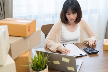 Güzel Asyalı kadın evde çalışıyor. İnternette sarı kutu ve dizüstü bilgisayarla çalışıyor. Müşterilerden isim siparişleri alıyor.