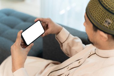 İslami genç adam oturma odasındaki kanepede oturuyor. Mutlu Arap adam cep telefonunu boş ekranla gösteriyor. Boş beyaz telefon ekranı gösteren mutlu Asyalı Müslüman adam. 
