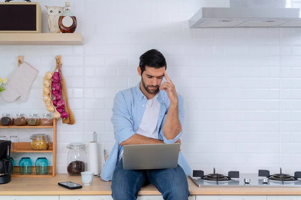 Handsome man using a laptop pc in the kitchen, concentrate and thinking with him job. Serious business man working at home having problem with job thinking to solve problem