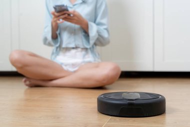Otonom robot daireyi temizliyor, genç bayanın boş vakti var. Kadın dinlenirken robotik süpürge odayı temizliyor. Uzaktan kumandayla vakumu kontrol eden kadın.