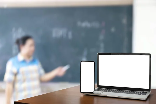 フロントビュー木製のテーブルの上に黒いデジタルノートパソコンとスマートフォンのブランク画面 教室の前に立つ先生 教育用ソフトウェアウェブサイト技術広告コンセプト — ストック写真