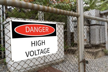 Dikenli tel örgülerin arkasında yüksek voltajlı trafo alt istasyonu ve Tehlike Yüksek Voltaj işareti var. Büyük kırmızı, beyaz ve siyah bir tehlike bu merkezden izinsiz geçenleri uyarıyor..