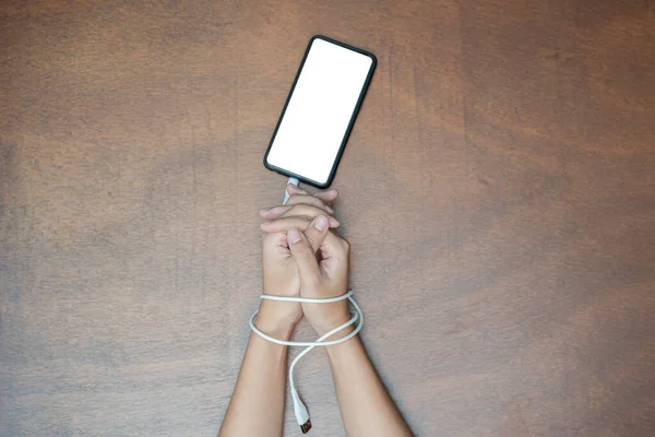 Woman hands trapped and wrapped on wrists with mobile phone cable as handcuffs in smart phone networking and communication technology addiction concept
