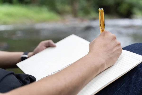 Young man takes notes in a notebook on nature. Man sits by a tree with his back to him, a pencil and a notebook in his hands, takes notes