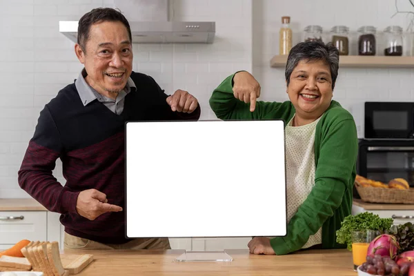 A retired couple shows off an empty laptop screen in the kitchen. Enjoy a relaxing retirement together. Free space for advertising communications.