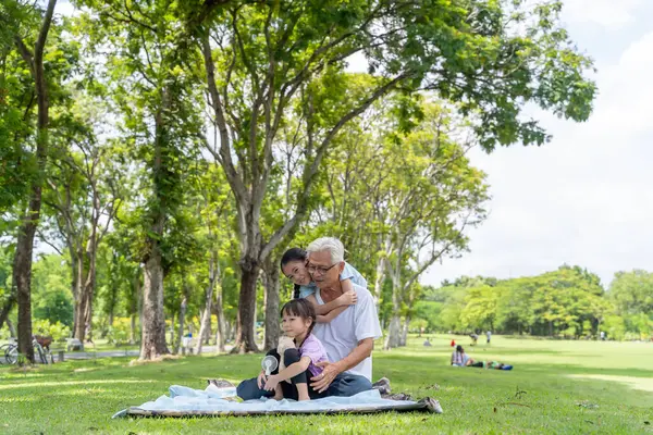 Two Asian granddaughters with grandpa and grandma sitting and talking on the grass outdoors in the park during the day. The age difference is a lot of warm love.