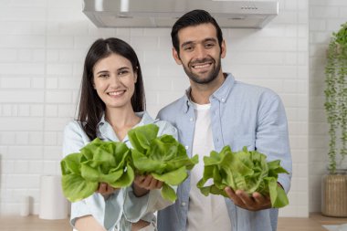 Genç çift yemek hazırlıyor, mutfakta sebze tutuyor, taze yeşil salata, ilaçsız. Yeni evlilerin sağlıklı yaşam tarzı, evde yemek pişirmek.