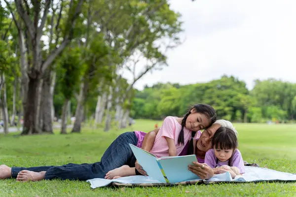 Happy family read books together and lying on green grass in public park. Little girl kid learning with grandfather in outdoors garden. Education and family lifestyle.