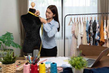 Asian mature female costume designer measure shape size of dressing puppet in a studio and works with clothing, fashion design collection, professional boutique tailor, dressmaker SME entrepreneur.