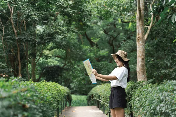 Woman with hat holding a map in the forest. Happy woman hiking and looking at map outdoors in nature. Travelers explore the mountainous forest map.