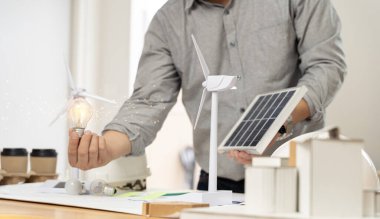 Close-up at a light bulb, An engineer sits holding a light bulb and model of wind turbine. with solar panels and model house on the table. To design use of renewable energy with wind and solar energy.