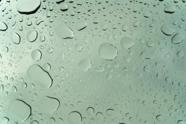 Üzerinde yağmur damlaları olan bir pencere. Yağmur damlaları pencerenin her tarafına saçılmış bulanık ve bulanık bir etki yaratıyor. Sahne sakin ve huzurlu, yağmur damlaları hafifçe düşüyor gibi.