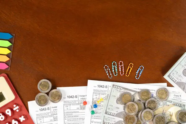 A table with a calculator, a stack of papers, and a pile of coins.  US tax form 1042 S with note and pen. tax form. law document usa. pink background. mathematics business concept.