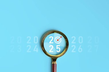 Moda araştırmaları, iş hedefleri ve mutlu yıllar 2025 kavramları. Altın büyüteçin içinde hedef simgesi olan büyük beyaz 2025 yılı numarası mavi arka planda, dikey biçimde elle tutuluyor..