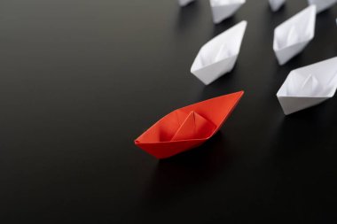 Bir grup beyaz kağıttan gemi tek yönde ve bir kırmızı kağıttan gemi siyah arka planda farklı yönleri gösteriyor. Yenilikçi çözüm kavramı için iş.