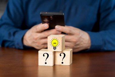 Yaratıcı fikir ve yenilik kavramını ele alalım. El, ampul ikonu ve soru işareti sembollü tahta küp bloğu seçti. Masada akıllı telefon kullanan bir adam yeni şeyler arıyor.