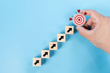 Hedef tahtası ve mavi arka planda ok olan tahta bloğu tutan bir adam. İş başarısı, hedef hedef ve proje ilerleme geçmişi, büyüme değeri, mali konsept