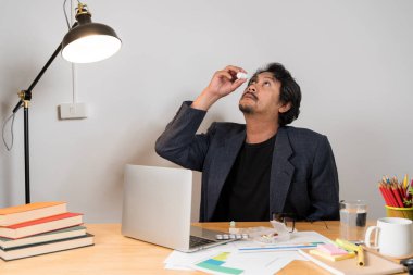 Aşırı çalışma ve hastalıktan gözüne ilaç damlatan genç Asyalı iş adamı. İş yerinin arkasındaki ofiste..