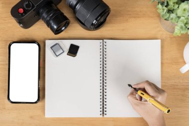 Bir insan kalemle bir tablete yazıyor. Ekran maketi olan bir telefon, kahve bardağı dolusu yazı malzemesi, kalemler, beyaz tahta masa arkaplanındaki bir not defteri. Çalışma alanının üst görünümü, alanı kopyala.