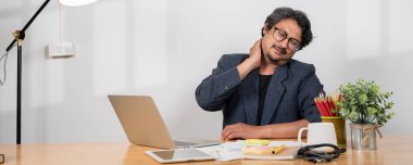 Yorgun, rahatsız Asyalı bir işadamı ofis sendromundan muzdarip bir bilgisayarın önünde masasında oturuyor. Rahatsızlığını hafifletmek için boynuna ve omzuna masaj yapıyor. sağlık hizmeti kavramı