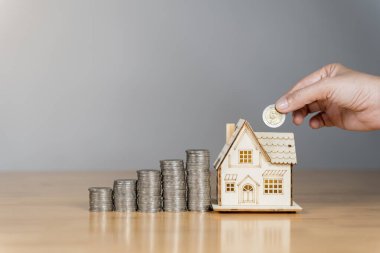 Adam cam kavanoza bozuk para ve yanında tahta bir ev modeliyle birlikte para atıyor. Gelecekteki konut yatırımları ve mali istikrar için para biriktirme kavramını vurguluyor