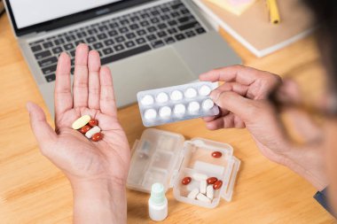 Genç adam evde dizüstü bilgisayar kullanırken ilaç alıyor. Hasta, hasta, çok çalışan. Elinde ilaç şişesi olan bir erkeğin eline ilaç dökerken yakın plan fotoğrafı..