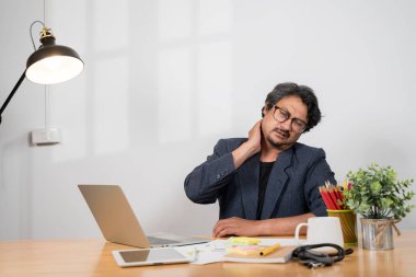 Sağlık sigortası kavramı. Yorgun, rahatsız Asyalı bir işadamı, ofis sendromundan muzdarip bir bilgisayarın önünde masasında oturuyor. Rahatsızlığını hafifletmek için boynuna ve omzuna masaj yapıyor..