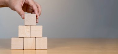 Bir el, bloklardan yapılmış bir piramit yapısının tepesine ahşap bir blok yerleştirir. Stratejiyi, planlamayı ya da inşaatı simgeler..