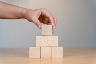 Bir el, bloklardan yapılmış bir piramit yapısının tepesine ahşap bir blok yerleştirir. Stratejiyi, planlamayı ya da inşaatı simgeler..