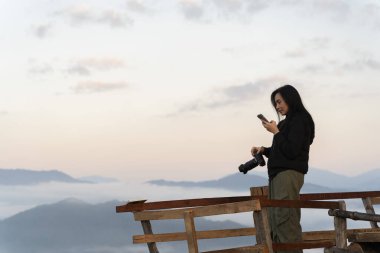 Güneş doğarken sisli dağlara tepeden bakan ahşap perspektifte akıllı telefon kullanan Asyalı bir kadın. Doğanın güzelliğini yakalamak ve anı paylaşmak. Seyahat ve bağlantı kavramı.