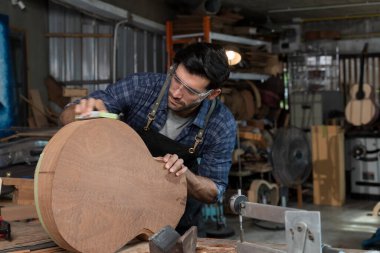 Erkek marangoz atölyede ahşap bir gitarın vücudunu zımparalıyor. Güvenlik gözlüğü ve önlüğü takıyor, işçiliği, ahşap işçiliğini, el yapımı müzik aletlerini ve profesyonel becerileri temsil ediyor..