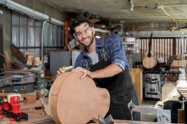 Ahşap bir atölyede gülümserken tahta gitar gövdesini zımparalayan kendinden emin bir erkek marangoz. Koruyucu gözlük ve önlük takmak, zanaatkarlık, tutku ve iş gücü göstermek.