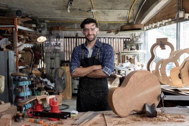Atölyede kolları çaprazlanmış, gitar parçaları ve aletlerle çevrili gülümseyen Luthier. Sanatçılık, ahşap işçiliği, el yapımı enstrüman yapımı ve yaratıcı stüdyo çalışma alanı kavramı.