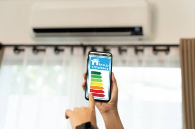 Elde tutulan akıllı telefon modern ev arka planında klimalı renkli enerji verimlilik tablosunu gösteriyor, çevre dostu teknoloji ve elektrik tasarrufu farkındalığını temsil ediyor.