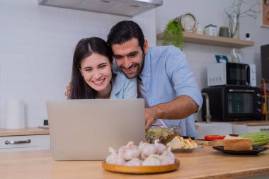 Modern mutfakta dizüstü bilgisayar ekranına bakan genç bir çift. Taze sebze ve malzemelerle çevrili. Gülümsüyorlar ve birlikte vakit geçiriyorlar. Yaşam tarzı, yemek pişirme ve aşk kavramı