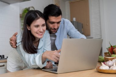 Mutfakta dizüstü bilgisayara bakıp gülümseyen ve sıcacık bir anın tadını çıkaran mutlu çift. Konforlu ev ortamında dijital yaşam tarzı ve modern teknolojiyi paylaşmak.