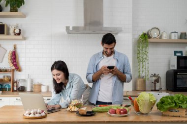 Genç çift mutfakta birden fazla iş yapıyor, kadın dizüstü bilgisayarda çalışırken gülümsüyor ve erkek akıllı telefon kullanıyor. Taze yiyecek ve sebzelerle çevrili, rahat bir evde modern dijital yaşam tarzının tadını çıkarıyor..