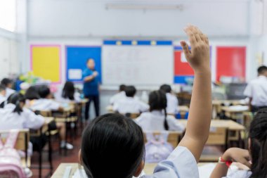 Öğrencilerin sıralarda oturduğu ilköğretim sınıfı sahnesi, bir çocuk katılmak için elini kaldırırken, öğretmen ön tarafta açıklıyor. Parlak ve ilgi çekici okul ortamı öğrenme ve etkileşime odaklandı.