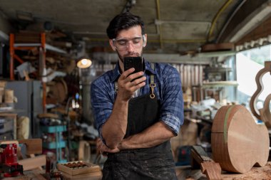 Güvenlik gözlüğü ve önlük takan genç bir marangoz atölyesinde akıllı telefona bakıyor. Modern zanaatkârlık kavramı, marangozluk ve yaratıcı endüstride teknoloji kullanımı