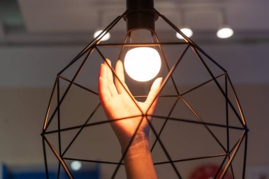 İnsan elinin yakın konum sıcak ampul dokunuyor dekoratif geometrik kablo abajur içinde sıcak modern iç oda. Enerji kavramı, sıcaklık, yaratıcılık ve ilham..