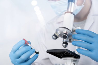 Laboratuvarda kan örneğini mikroskop altında inceleyen bilim adamı, eldiven ve maske takıyor, tıbbi araştırmaları, sağlık hizmetlerini, mikrobiyoloji çalışmalarını, yenilikleri ve bilimsel ilerlemeyi temsil ediyor.