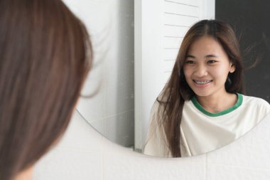 Banyoda diş tellerini gösteren genç bir kadın aynada gülümsüyor. Ortodontik bakım, sağlıklı dişler, kendine güven, ağız temizliği, diş güzelliği ve mutlu yaşam tarzı kavramı..