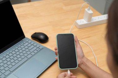 Akıllı telefon tutan ve şarj kablosunu ahşap bir ofis masasında dizüstü bilgisayarın ve farenin yanına bağlayan biri, modern teknoloji ve iş yaşam tarzını minimum çalışma alanı ortamında gösteriyor.