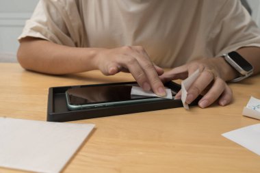 Ekran koruyucusu takmadan önce ıslak mendili akıllı telefon ekranını temizlemek için kullanana yakın çekim. Elleri temiz bez tutarken, ahşap bir masada alet hazırlarken. Teknoloji hijyeni kavramı.