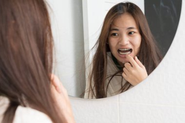 Uzun saçlı Asyalı kadın aynaya bakıyor ve diş tellerini işaret ediyor. Günlük banyoda diş bakımı, ağız temizliği, ortodonti, diş düzenleme ve sağlık rutini kavramı.