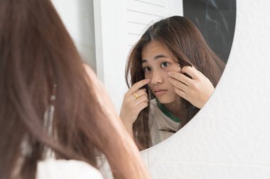Asyalı bir kadın banyo aynasında yüzünü inceliyor, derisine endişe ile dokunuyor. Cilt bakımı, sivilce, leke, siyah halkalar, güzellik rutini veya cilt sağlığı sorunu ve yüz bakımı farkındalığı kavramı.