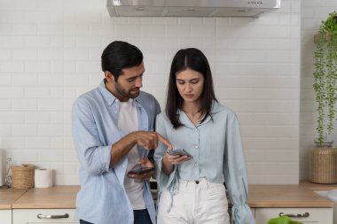 Modern ev mutfağında birlikte duran genç çift akıllı telefon kullanıyor, kadın bakarken, adam mobil ekranı işaret ediyor, iletişim, dijital yaşam tarzı, teknoloji kullanımı, çevrimiçi bağlantı.