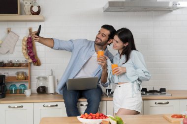 Modern mutfaktaki genç çift akıllı telefonlarıyla selfie çekerken gülümsüyor, ellerinde portakal suyu bardakları, mutluluk, yaşam tarzı, ilişki, günlük ev sahneleri, neşeli ve pozitif anlar.