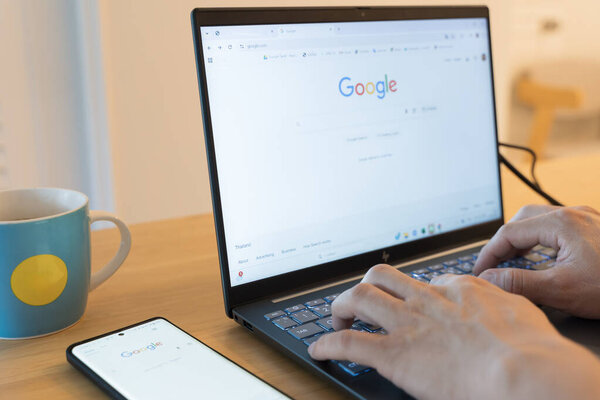 5 октября 2025 Бангкок, Таиланд: Крупный план руки человека, печатающего на клавиатуре ноутбука, выполняющего поиск на домашней странице Google. Смартфон и кофейная кружка сидят на деревянном столе.