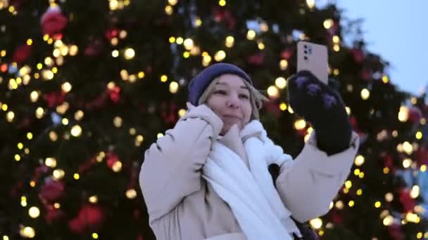 街の広場にある美しいイルミネーションクリスマスツリーの近くに立って電話で話す楽しい女性 — ストック動画